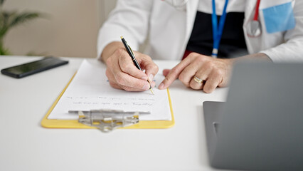 Middle age man with grey hair doctor using laptop writing on clipboard at clinic