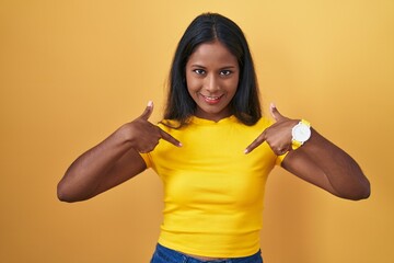 Young indian woman standing over yellow background looking confident with smile on face, pointing oneself with fingers proud and happy.