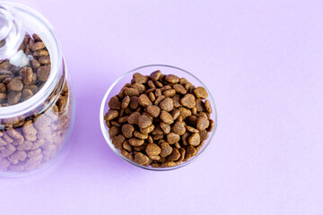 Dry pet food in a glass jar and bowl close-up on a lilac background