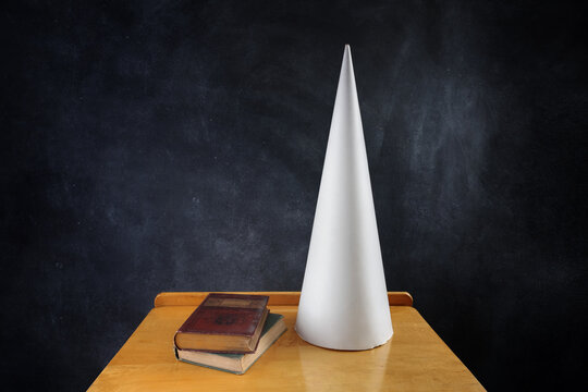 Dunce cap on a desk with books, negative words written on the chalkboard
