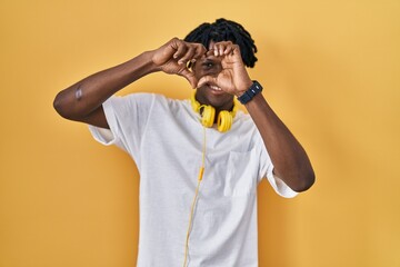 Young african man with dreadlocks standing over yellow background doing heart shape with hand and fingers smiling looking through sign