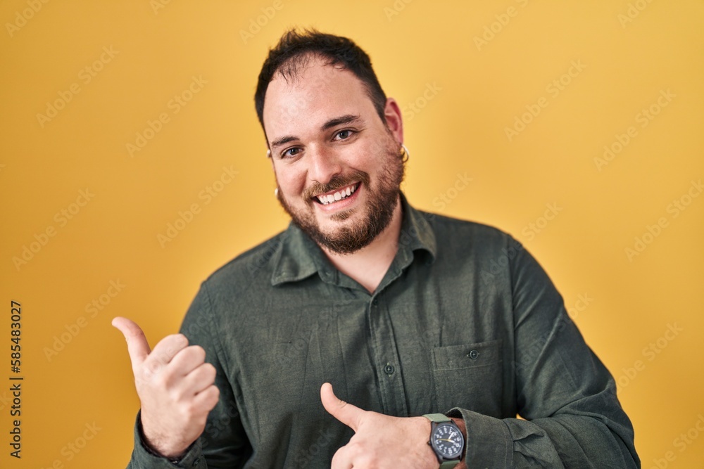 Wall mural plus size hispanic man with beard standing over yellow background pointing to the back behind with h