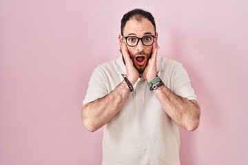Plus size hispanic man with beard standing over pink background afraid and shocked, surprise and amazed expression with hands on face