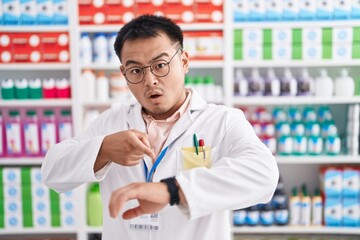 Chinese young man working at pharmacy drugstore in hurry pointing to watch time, impatience, upset and angry for deadline delay