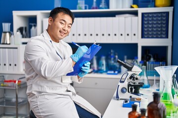 Young chinese man wearing scientist uniform writing on clipboard at laboratory