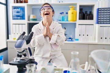 Hispanic girl with down syndrome working at scientist laboratory begging and praying with hands together with hope expression on face very emotional and worried. begging.