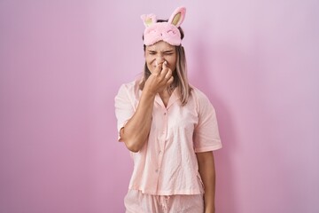 Blonde caucasian woman wearing sleep mask and pajama smelling something stinky and disgusting,...