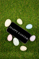 Phone with Easter greetings on a black background of easter eggs with in on green grass. Space for text. Happy Easter! Smartphone mockup, top view