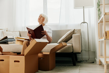 elderly woman sits on a sofa at home with boxes. collecting things with memories albums with photos and photo frames moving to a new place cleaning things and a happy smile.