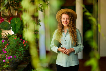 Young woman stands in garden holding gardening tool in her hands smiles and looks at camera. Junior female gardener with a trowel posing in green orchard. Farming and gardening concept