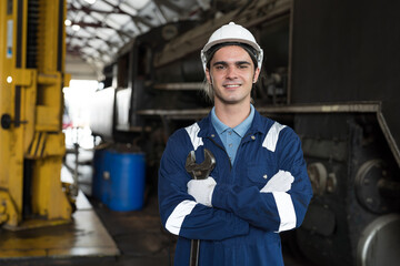 Male engineer worker working in industry factory, standing with crossed arms, smile and holding industrial spanner, wearing safety uniform, helmet. Male technician worker at work in locomotive garage