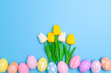 Happy Easter holiday greeting card design concept. Colorful Easter Eggs and spring flowers on blue background. Flat lay, top view, copy space.
