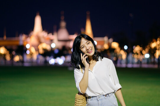 Portrait young beautiful asian woman smiling and using smartphone while travel at Temple of the Emerald Buddha or Wat Phra Kaew in night view point, Bangkok, Thailand.