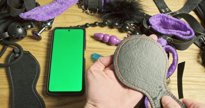 Man's hands check the bdsm spatula for spanking. Smartphone with green screen, sex toys lie on the table. Top view, close-up.
