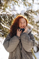 View of a cute smiling woman in a gray down jacket. A girl with red hair walks in the park on a sunny winter morning.