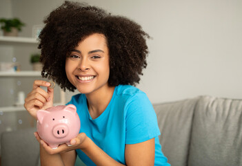 Fototapeta na wymiar Smiling young curly african american woman put coin into piggy bank in living room interior, close up