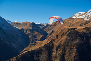 a paraglider with a white wing is gliding against the backdrop of sharp mountain peaks with snowy tops against the backdrop of a beautiful clear blue sky