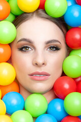 Fashion, pop-art and make-up concept. Beautiful woman close-up studio portrait in colorful balls background. Model's head surrounded with various colors plastic balls. Girl looking at camera