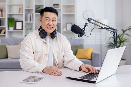 Portrait of blogger at home, Asian man smiling and looking at camera, man sitting at table with laptop in living room, recording audio podcasts and doing online radio.