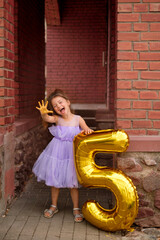 A girl in a purple dress laughing merrily showing five fingers on her hand. A balloon in the shape...