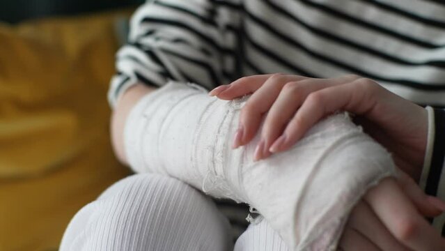 Close-up cropped shot of unrecognizable young woman with broken hand wrapped in white plaster bandage, suffering from pain, gently massaging injured forearm, sitting at home. Shooting in slow motion.