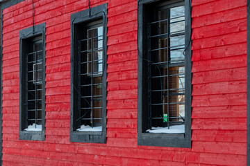 Fototapeta na wymiar Three closed double hung glass windows with black trim on a vibrant red wooden clapboard building. There's white snow on the window sill of each window. The windows have metal bars covering the glass 