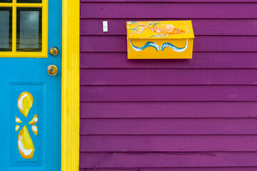 A yellow mailbox with decorative painted ribbon hangs on a purple exterior wall of a house. The...