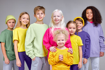 cheerful kids in fashion stylish clothes standing in row behind little girl with fluffy curly fair hair, children support little kid, help her