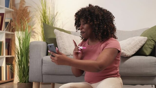 Pregnancy excitement. Mobile call. Happy woman. Cheerful black female showing maternity test to smartphone online meeting sitting floor light room interior.