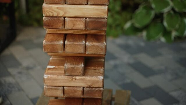 Unrecognizable adult man with hairy hands and gold wristwatch in shirt takes out wooden block from jenga tower, close-up. Man in leather jacket approaches wooden game. People play logic game outdoor.