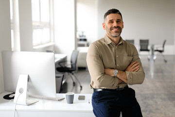 Successful middle aged male entrepreneur sitting with arms crossed at desk in office, smiling at...