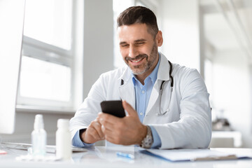 Smiling male doctor in medical uniform sitting at desk in clinic, looking at phone screen,...