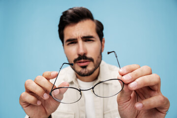 Close-up portrait of a brunette man looking through glasses that he holds in his hands, eye problems, glasses for vision farsightedness and nearsightedness, on a blue background, copy space 