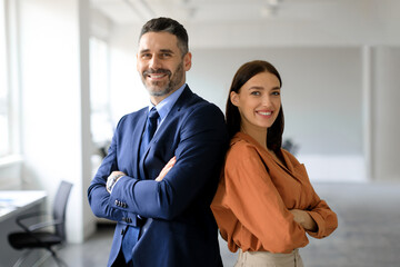Two successful businesspeople standing back to back with arms crossed and smiling at camera, posing in modern office - obrazy, fototapety, plakaty