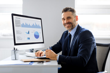 Portrait of happy businessman working on computer in office and smiling at camera, monitor with...