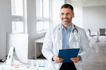 Portrait of middle aged male doctor holding clipboard, looking aside and smiling, physician gp...