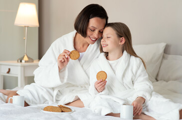 Smiling caucasian small girl and young woman in bathrobe sit on white bed with cups, drink tea and eat cookies