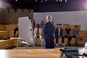 Woman University Professor Holds Lecture to Classroom Full of Multi Ethnic Students. Prominent Lecturer Teaching Bright Young People. Rear View, Focus On Short-haired Speaker Woman In Formal Wear
