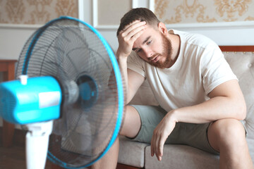 Young bearded man using electric fan at home, sitting on couch cooling off during hot weather,...