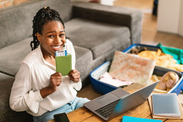 Excited african american woman with passport and tickets smiling at camera, sitting with laptop near unpacked suitcase
