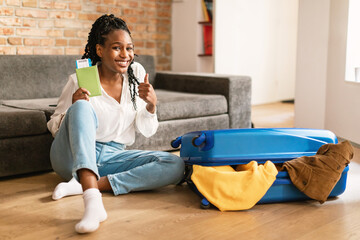 Happy african american female tourist sitting near suitcase, holding passport with tickets and...