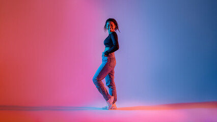 Fototapeta na wymiar Full length of stylish african american woman posing in neon light in studio and smiling at camera
