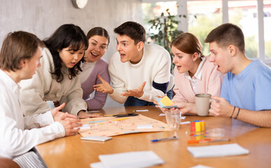 Cheerful adult students playing board game excitedly sitting around the table
