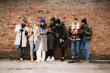 Mixed race group of stylish friends standing against a wall in outdoors smiling and using mobile phone.