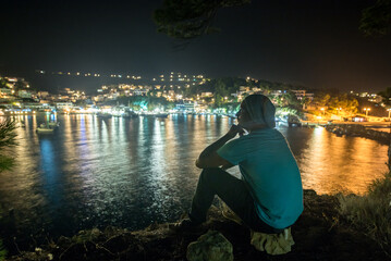 Mann mit Strickmütze genießt Ausblick auf nächtlich beleuchtetes Fischerdorf am Mittelmeer