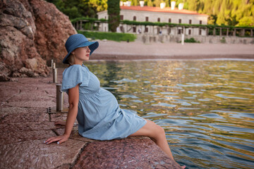 Pregnant woman traveler in denim dress, blue straw hat sits by the sea, dangles legs in water