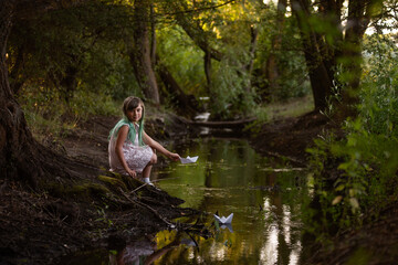 Fototapeta na wymiar Teenage girl with green hair in dress, stands on the river bank, launches white paper origami boat