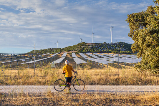 Castilla y Leon, Spain. Man from behind leaning on bicycle looking at wind power towers and solar farm in rural setting at sunset