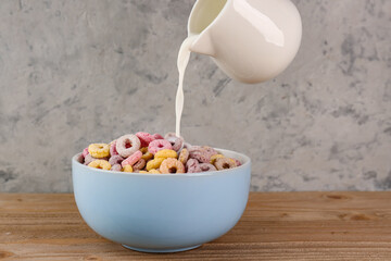 Pouring of milk from pitcher into bowl with colorful cereal rings on wooden table near grey grunge wall