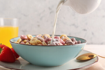Pouring of milk from pitcher into bowl with colorful cereal rings on table against white grunge...